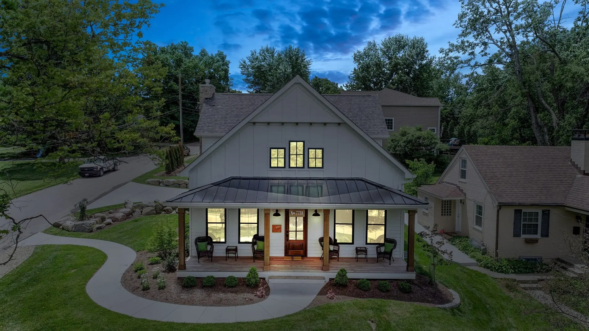 7731 Dones Ave. in Madeira from above at dusk: a grey farmhouse-style home with a deep front porch and every window glowing, a curving front walk and mature trees around it