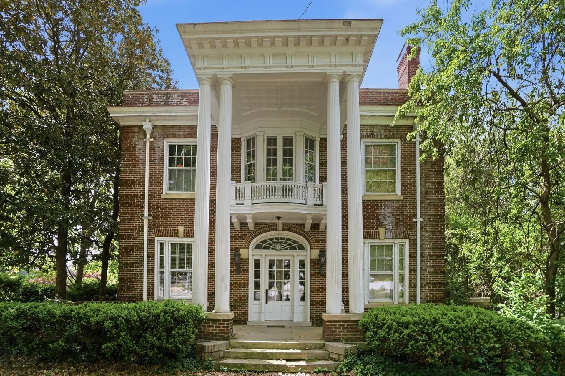 2925 Erie Ave. before restoration: a 1911 red-brick home with a two-story white-columned portico, a balcony over an arched front door and boxwood hedges along the front