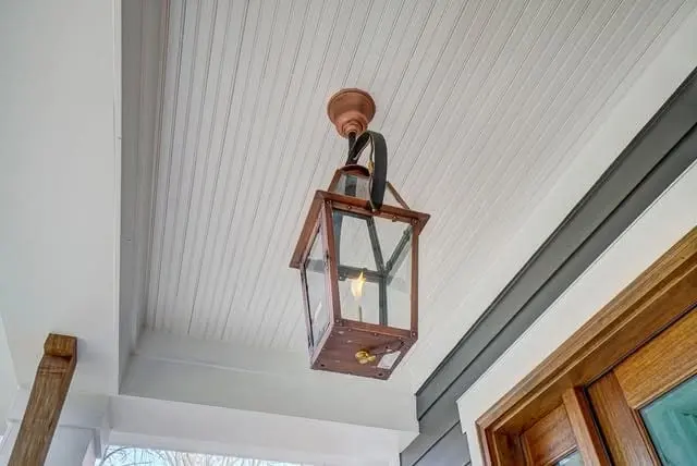A copper gas lantern hanging from a white beadboard porch ceiling beside a stained wood front door