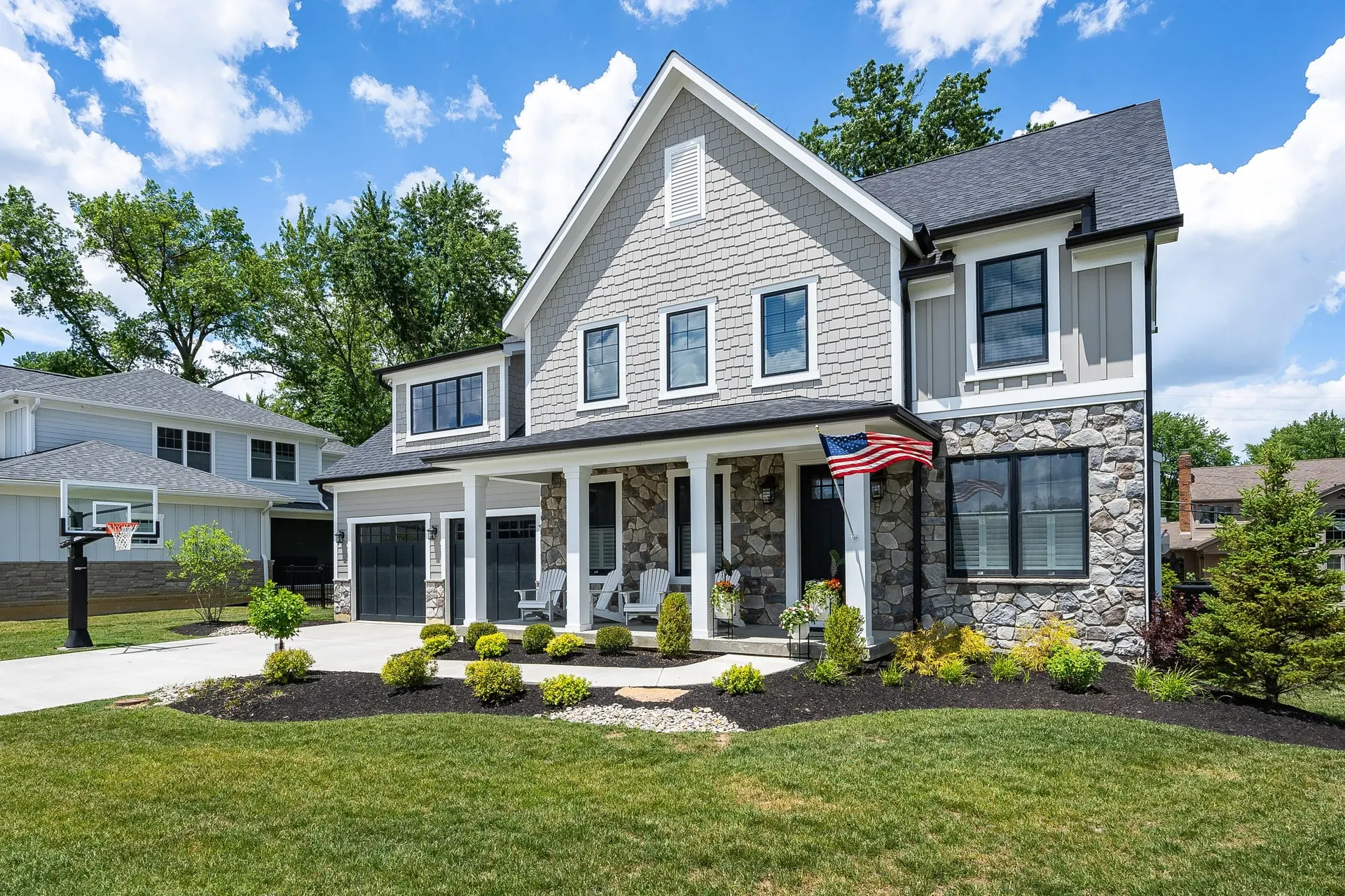 4890 Heitmeyer Lane: a grey two-story home with a stone-clad wing, black-framed windows and a long covered front porch flying an American flag