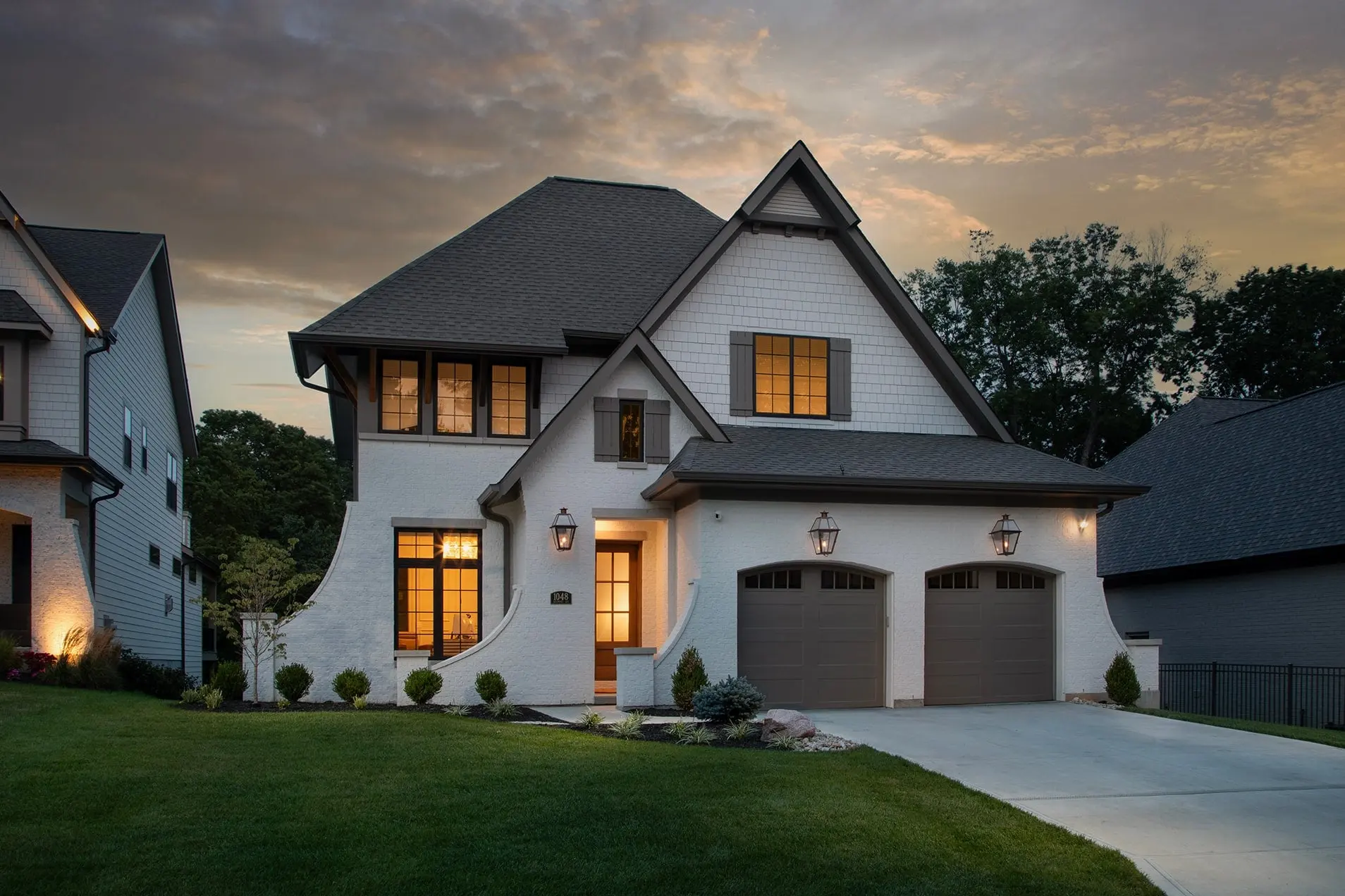 1048 Richwood at dusk: a white two-story home with a steep gable, a sweeping curved wall beside the front door, lit wall lanterns and glowing windows under a dramatic evening sky