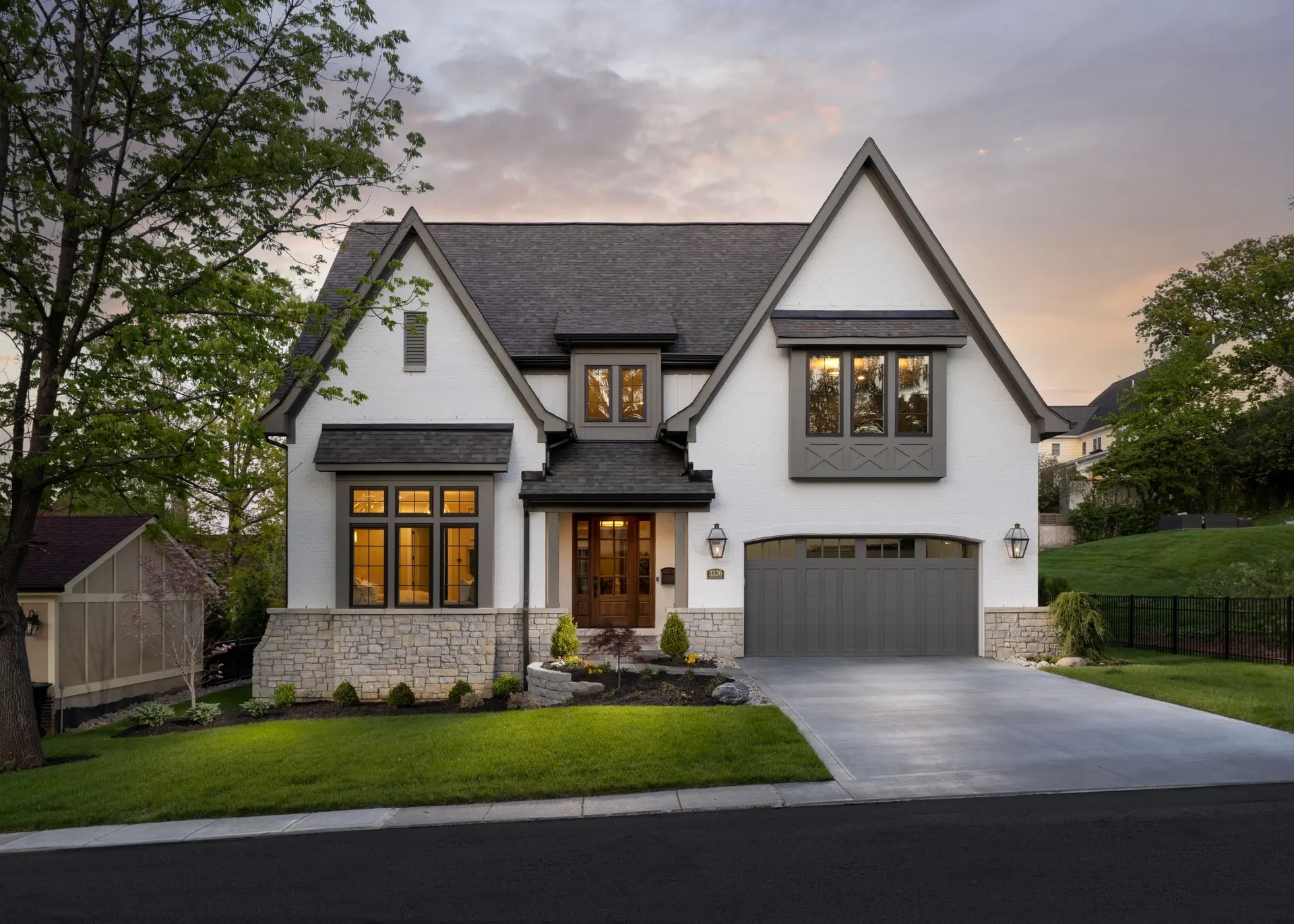 3326 Stettinius at dusk: a white Tudor-style home with steep gables, a stone base, a wood front door and every window glowing under a soft peach sky