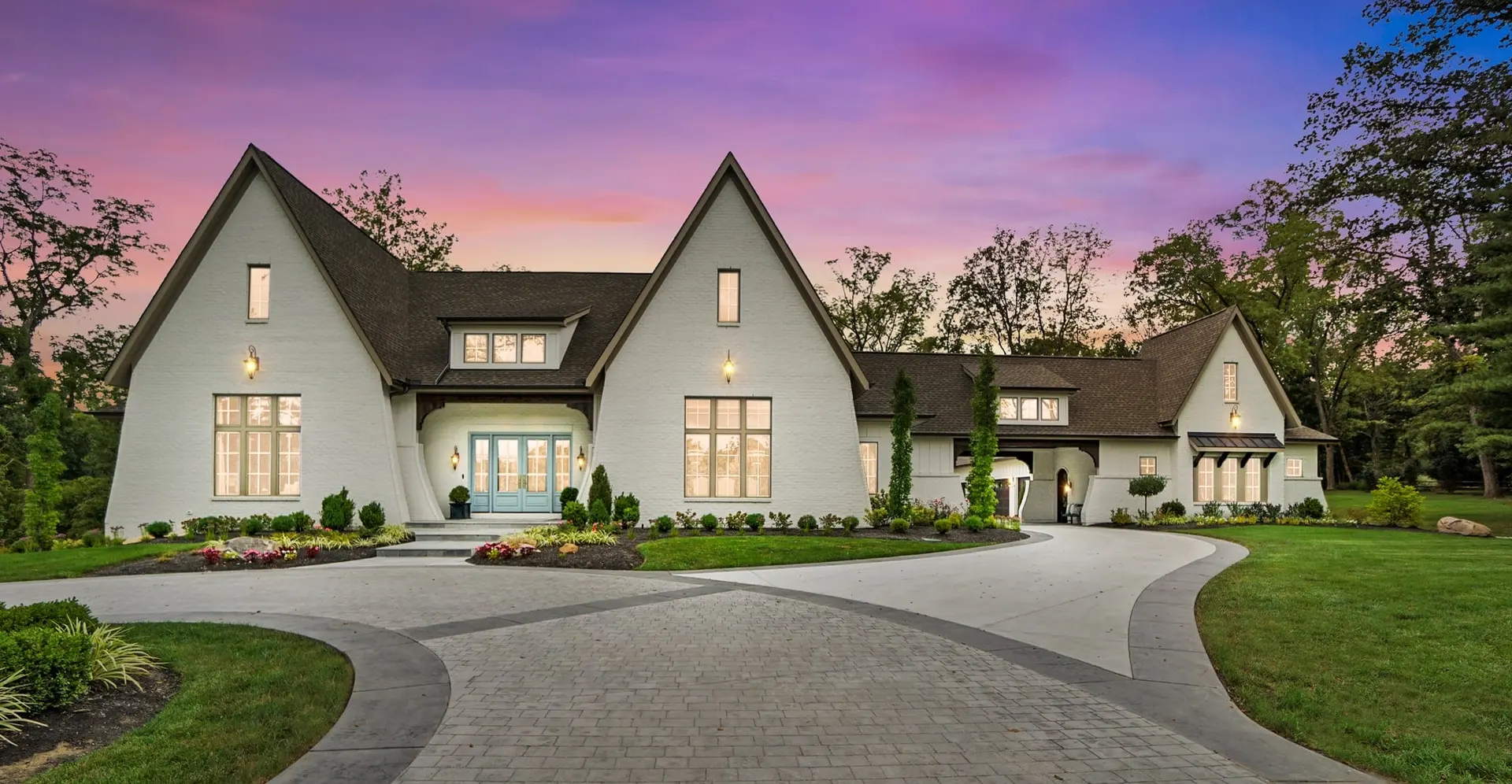 A long white Tudor-style home at twilight, with two steep front gables, warm lit windows and a curving paver driveway under a pink and violet sky