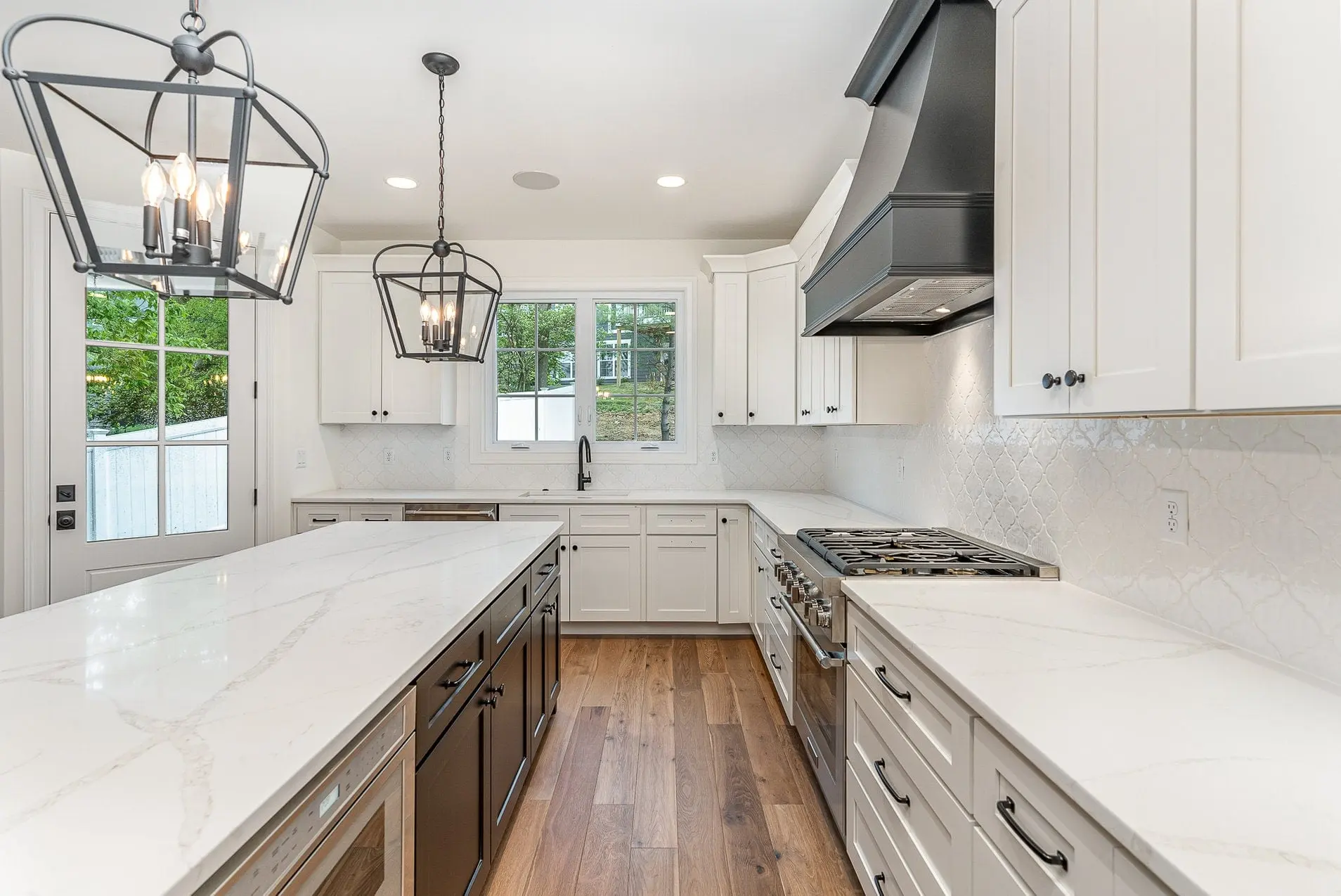 Two black iron lantern pendants over a long white quartz island with a dark base, white shaker cabinets, a charcoal range hood and wide-plank oak floors