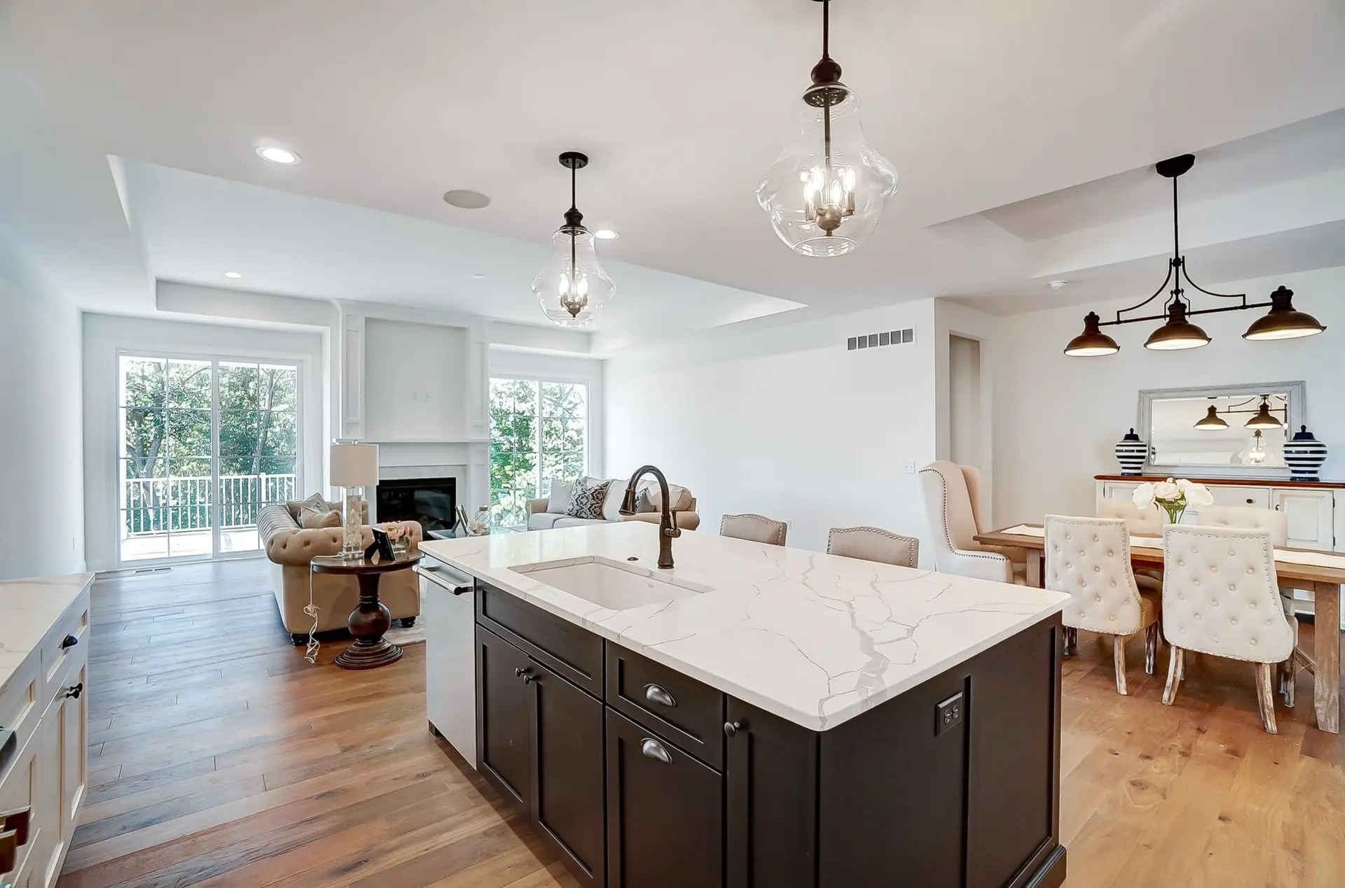 An open kitchen, dining and living space in a Laurel Crossing home: a dark island with a white quartz top under glass pendants, a dining table with tufted chairs and a fireplace beyond
