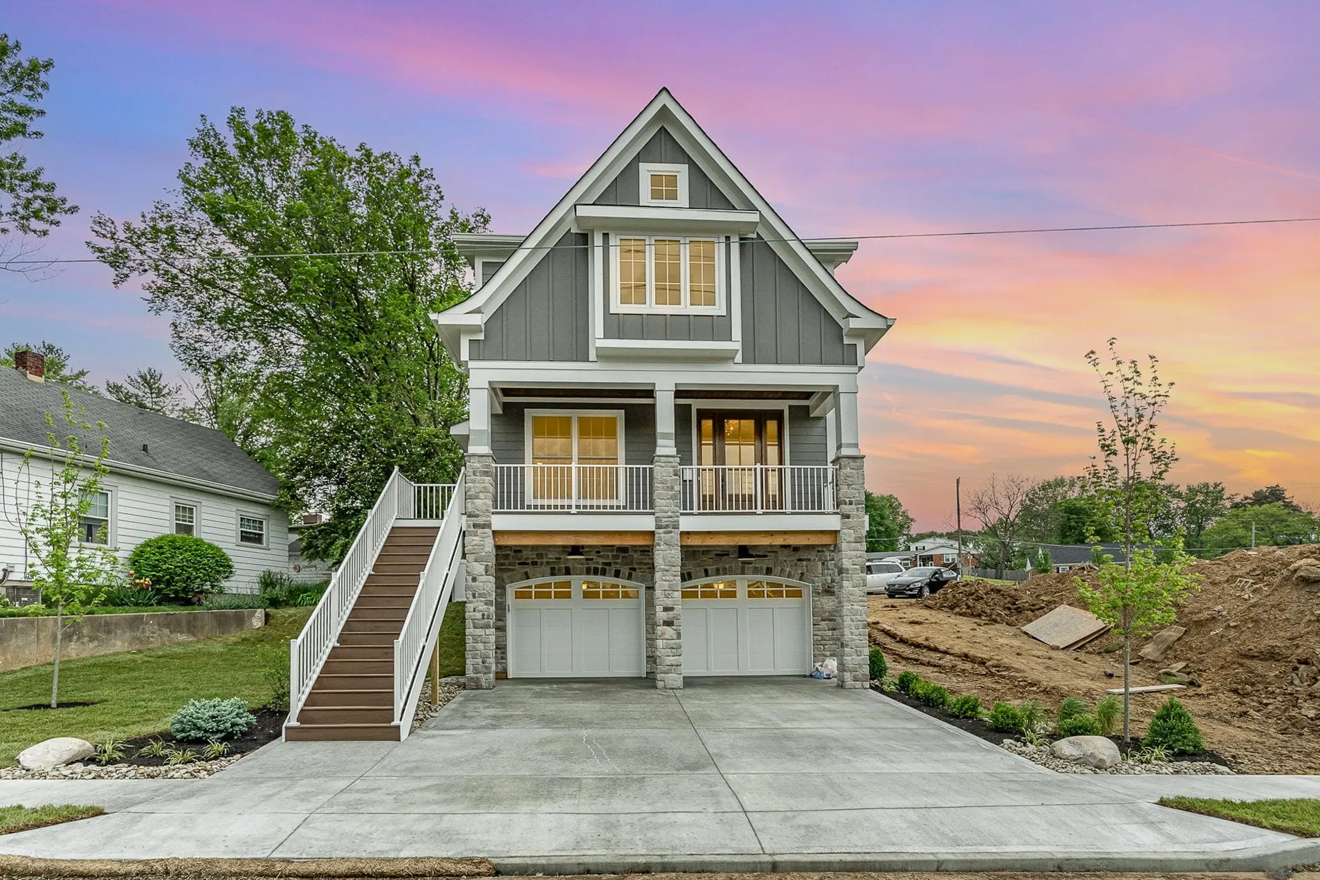A new Laurel Crossing home in Madeira at sunset: grey board-and-batten siding, a raised front porch with white railings over a stone two-car garage and an outdoor stair, under a pink and orange sky