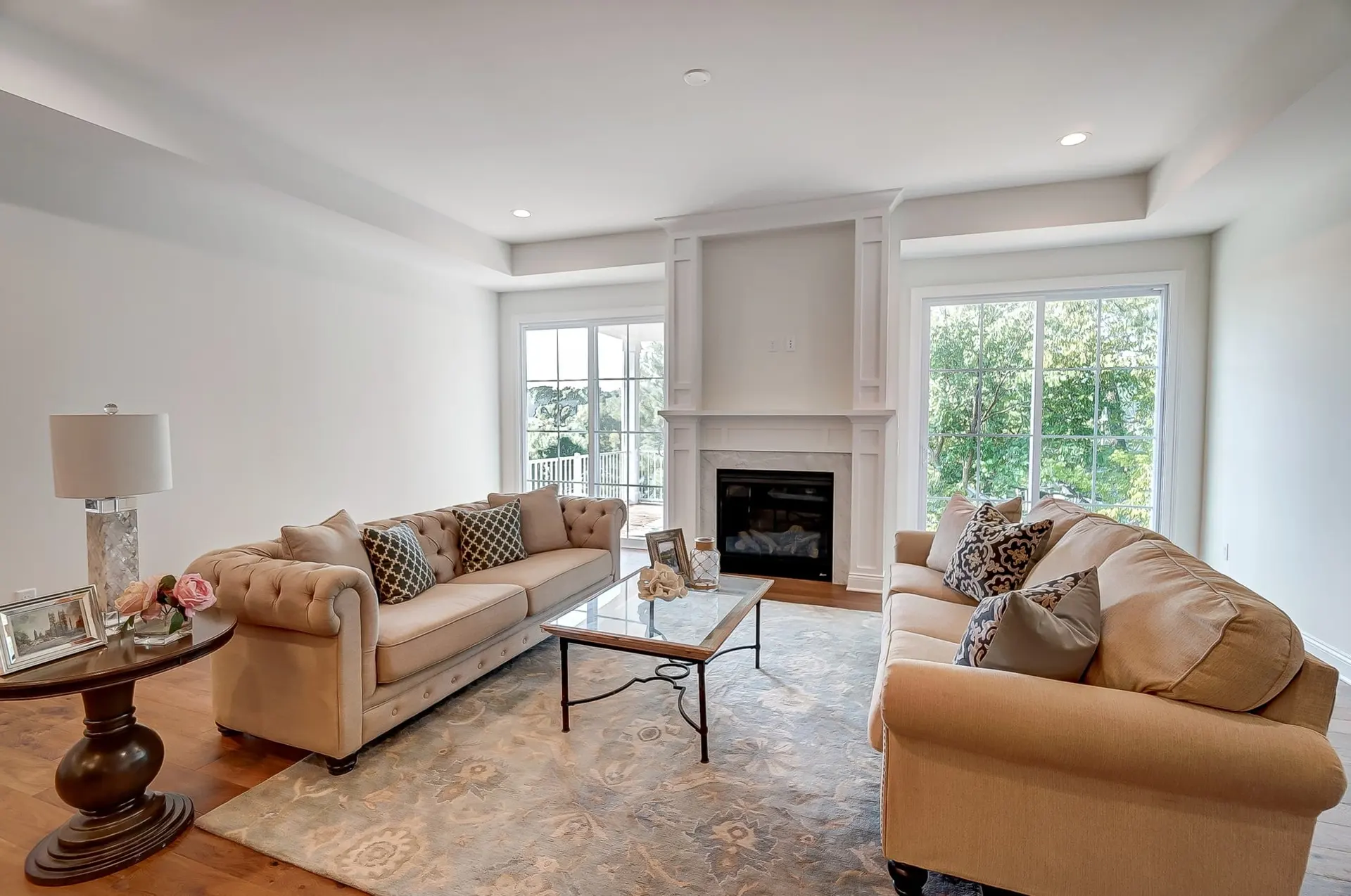 A staged living room with two tan sofas facing a fireplace, tall windows on both sides, a patterned rug and oak floors