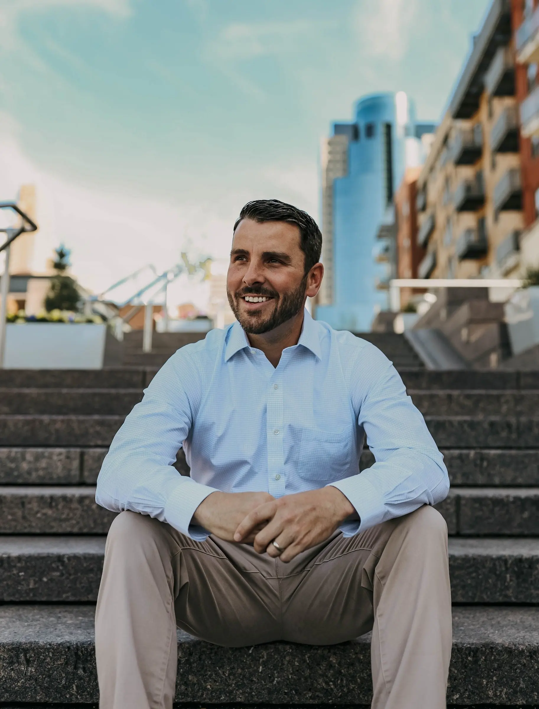 Matt Stanley, owner of Legendary Homes, smiling as he sits on wide stone steps in a light blue shirt, with city buildings behind him