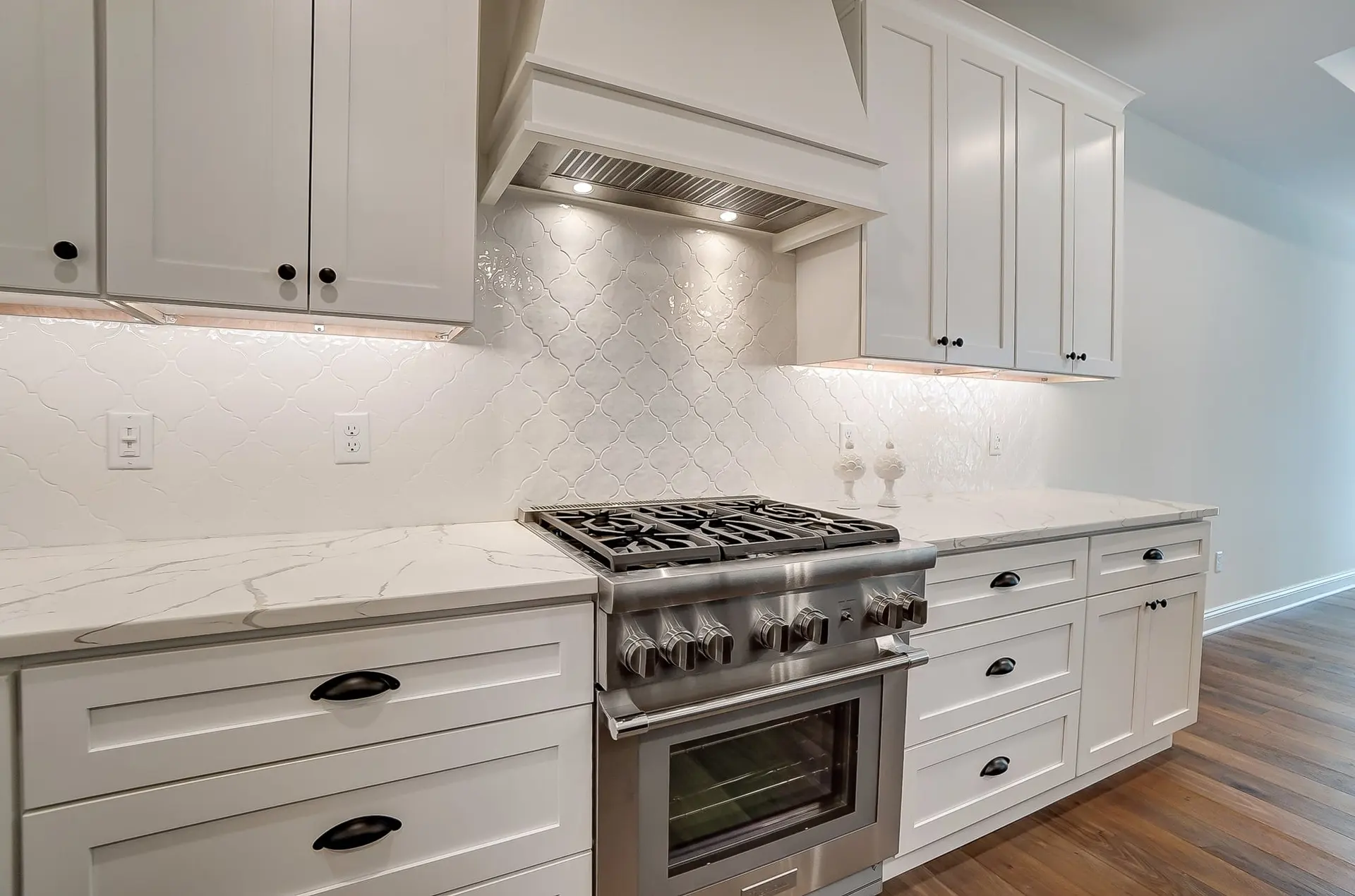 A stainless steel gas range between white shaker cabinets with black cup pulls, under a white hood, with a white arabesque tile backsplash and veined quartz counters