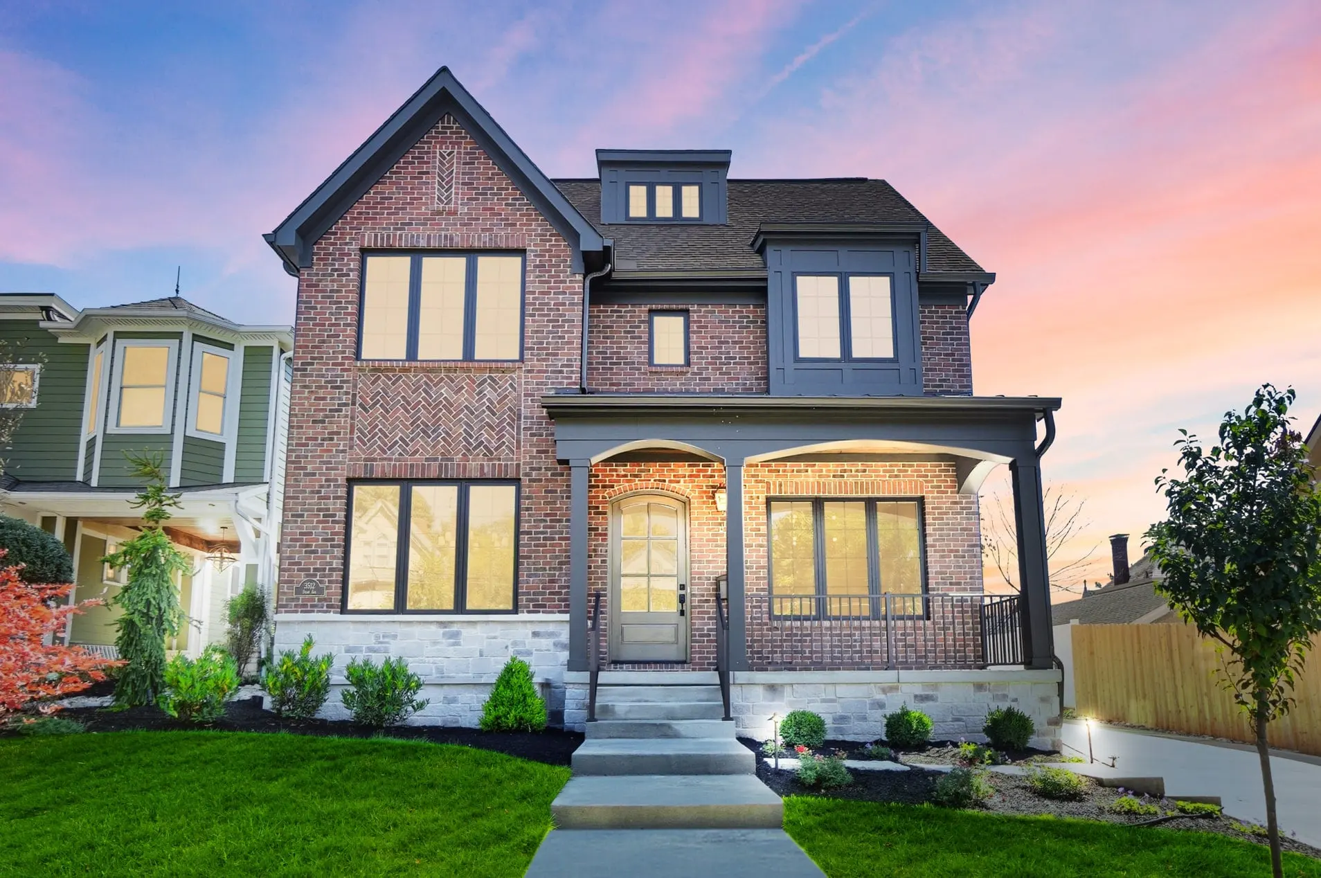3512 Shaw at sunset: a red-brick home with dark-trimmed windows, a herringbone brick panel and a lit arched front porch above stone steps, under a pink and blue sky