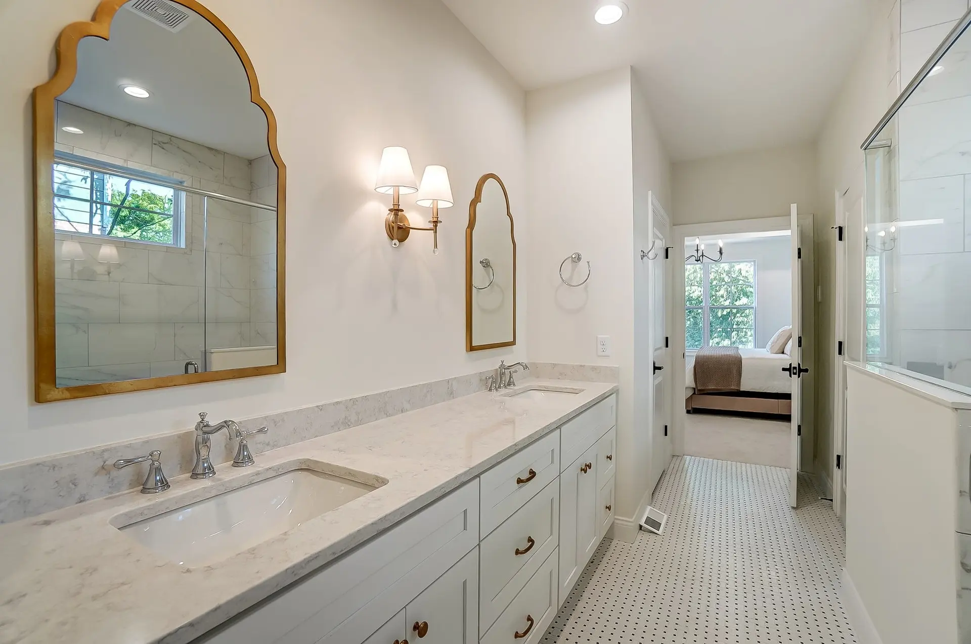 A long white bathroom vanity with a marble-look counter and brass pulls, two arched gold-framed mirrors with a brass double sconce between them, and a mosaic tile floor leading to a bedroom